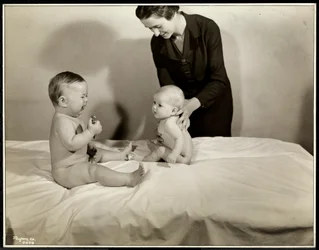 To babyer som sitter oppe i sengen, den ene blir støttet av en sykepleier, den andre holder en lekebil og gråter på Maternity Center Association i 70th Street East River, New York, 1935 (sølv gelatintrykk
